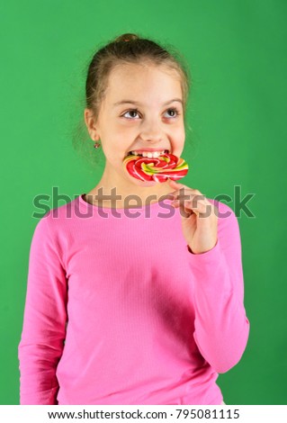 Child with hungry face poses with candy on green background. Lady holds round shaped lollipop. Happiness and dessert concept. Girl eats big colorful sweet caramel.