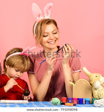Mother and daughter preparing for Easter. Easter celebration concept. Family members wearing cute bunny ears. Woman and kid with happy faces painting eggs on pink background.