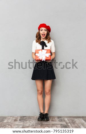 Full length portrait of an excited schoolgirl dressed in uniform holding gift box and looking at camera over gray wall background