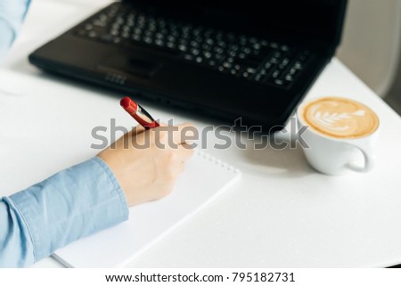 girl freelancer in a blue shirt writes in a notebook, on the desk is a black laptop and coffee