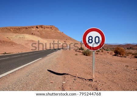 Road sign speed limit 80 km on the side of empty asphalt road through rocky desert, in the background hills, sunny day, blue sky