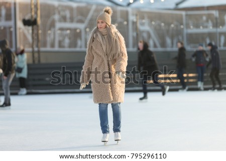 Full length portrait of young woman with blonde hair in fur coat, beige hat, scarf and blue jeans on ice rink, outdoors at sunny winter weather/ Weekends activities outdoor in cold weather/