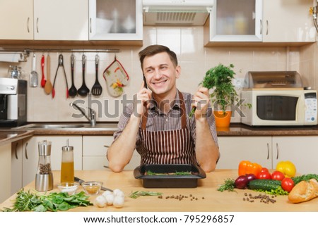Man in apron sitting at table with vegetables, dill, talking on mobile phone, cooking at home preparing meat stake from beef or lamb, in light kitchen with wooden surface, full of fancy kitchenware