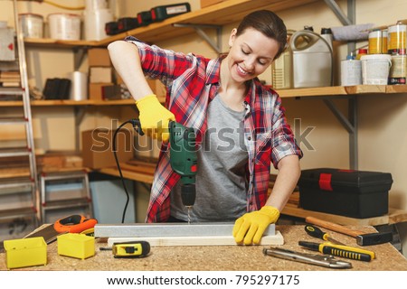 Beautiful caucasian young brown-hair woman in plaid shirt and gray T-shirt working in carpentry workshop at table place, drilling with drill holes in piece of iron and wood while making furniture
