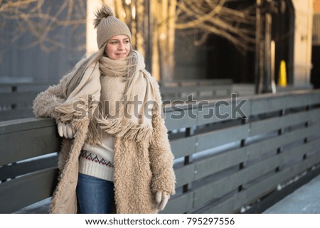 Full length portrait of young female with blonde hair in fur coat, beige hat, scarf and blue jeans posing alone and smiling on ice rink, outdoors at winter/ Weekends activities outdoor in cold weather