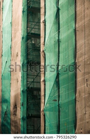 Scaffolding covered by thin layer of white and green flexible plastic textile, illuminated by warm afternoon light. Maintenance work on residential building in Berlin, Germany. Vertical closeup view.