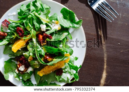 Overhead of arugula salad on white plate with candied nuts, orange segments, ricotta cheese and pepper on brown surface with silver fork.