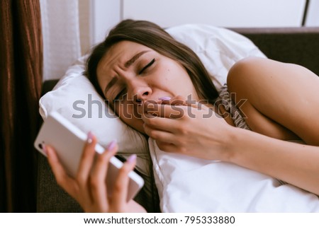 sleepy young girl lies in bed under a white blanket, yawns and looks at smartphone