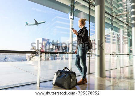 Young casual female traveler at airport, holding smart phone device, looking through the airport gate windows at planes on airport runway. Royalty-Free Stock Photo #795378952