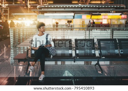 Lonely Brazilian girl is sitting inside of modern airport terminal on metal seats and waiting her flight; cute young African American female tourist is waiting for her train indoors of railway station