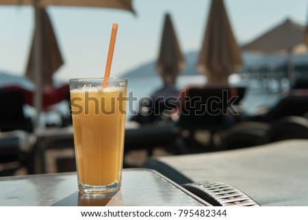 Glass of orange juice with straw on the background of blurred chaise lounges on the beach on sunny summer day, copyspace