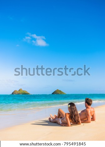 Beach vacation couple relaxing sunbathing on hawaiian tropical beach in Lanikai, Oahu, Hawaii, USA. American people on summer holidays sun tanning lying down on sand.