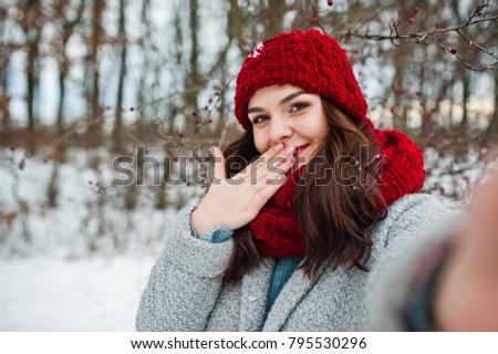 Portrait of gentle girl in gray coat , red hat and scarf near the branches of a snow-covered tree, holding camera for selfie.