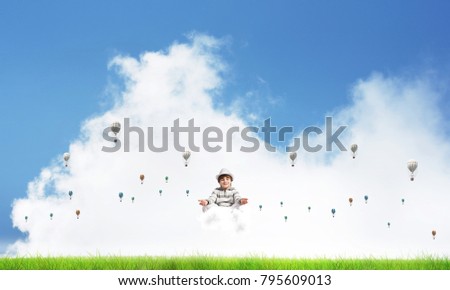 Young little boy keeping eyes closed and looking concentrated while meditating on cloud among flying aerostats with bright and beautiful landscape on background.