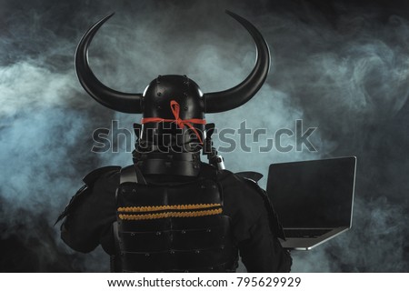 rear view of samurai in traditional armor with laptop on dark background with smoke Royalty-Free Stock Photo #795629929