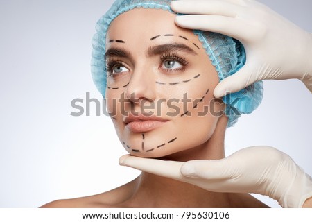 Beauty portrait of woman in blue cap looking aside, plastic surgery concept, studio. Hands in white gloves holding face of model with puncture lines, indoors Royalty-Free Stock Photo #795630106