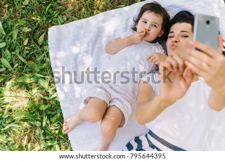 Overhead view of young blowing kiss mother taking self portrait with beautiful little girl daughter lying on white plaid on grass. Young family taking selfie outdoor. Motherhood and childhood concept