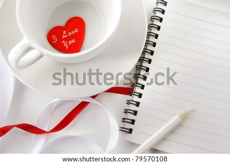 White tea cup with note-pad, pencil and red heart.