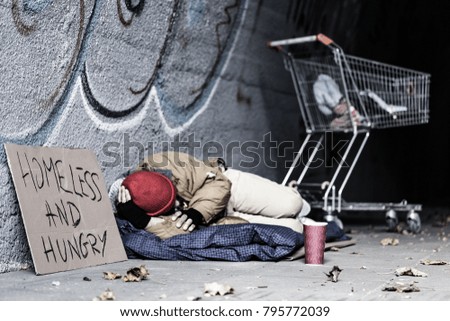 Dirty tramp lying on blanket next to a sign and trolley in the shelter of the city Royalty-Free Stock Photo #795772039