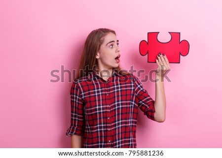 A young surprised girl holds a large puzzle on raised hand and looks at it. On a pink background.