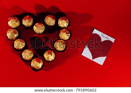 On a red background is a box in the shape of a heart and in it is candy in a Golden wrapper. Next lay a piece of paper with a cartoon heart