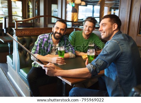st patricks day, leisure and technology concept - happy male friends drinking green beer and taking picture with smartphone selfie stick at bar or pub