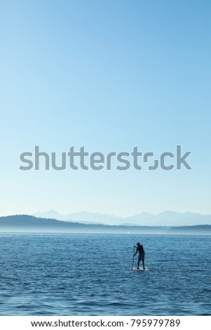 Silhouetted stand up paddle boarding man, digging in, on a calm and vibrant blue ocean, with mountains on the horizon