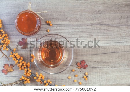 Autumn healthy hot drink concept. Branch of common sea buckthorn with berry, cup of tea, jar of jam on light wooden background. Toned photo with copy space for your text. Top view.