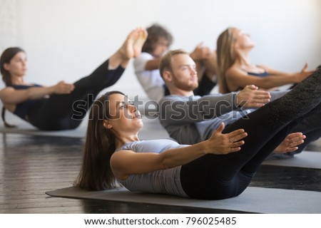 Group of young sporty people practicing fitness lesson with instructor, doing warming up exercise, boat pose, working out, students training in club, friends at fitness lesson, indoor, studio Royalty-Free Stock Photo #796025485