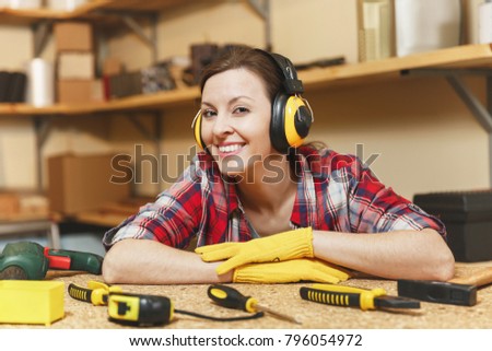 Pretty caucasian young brown-hair woman in plaid shirt, gray T-shirt, noise insulated headphones, yellow gloves working in carpentry workshop at wooden table place with piece of wood, different tools