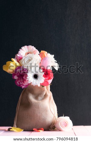 Canvas gift bag filled with multicolored roses, tulips and gerbera, on a pink table and a black background, surrounded by a rose and petals.
