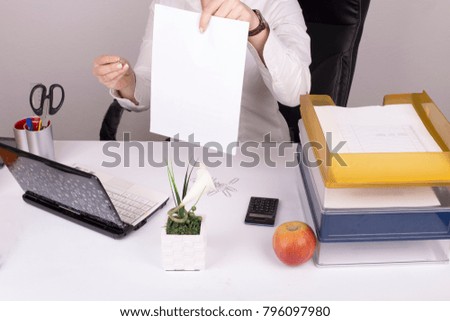 Notebook, pen holder, calculator, paper holder, an apple and plant standing on  desk.  Girl sitting in chair in background holds a piece of paper. 