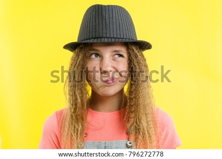 Pretty girl 12-13 years old blonde with curly hair in a hat, looks pensively aside, thinking about school. Facial expressions concept, yellow background