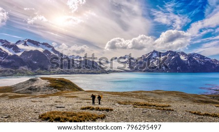Wide view of the beautiful nature landscape of South Georgia Island, with a dramatic sky, snow-capped mountains, a large saltwater bay, and barren foreground, including two unidentifiable hikers.  Royalty-Free Stock Photo #796295497