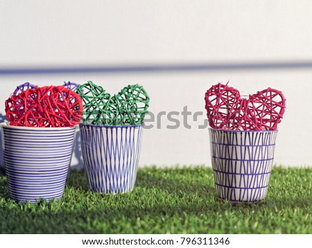 Three porcelain glasses are on the green grass. In glasses there are multi-colored wicker hearts. In the background there is a bright wall with a strip of shadow. Picture for Valentine's day or Easter