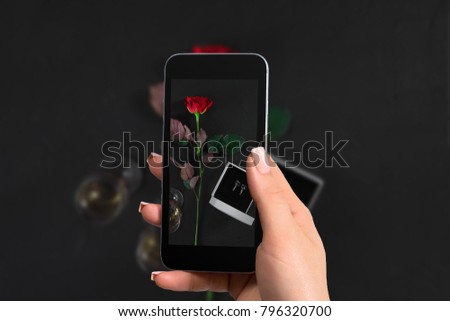 Female hand with a smartphone makes a photo one dark red rose and box with earrings on black background.