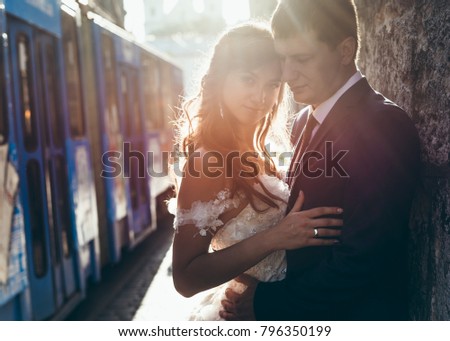 Sensitive portrait of the happy newlywed couple hugging and leaning on the wall at the background of the driving tram during the sunset.