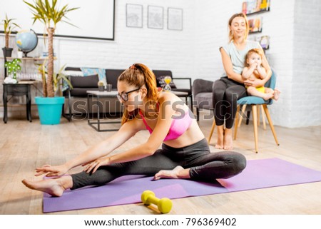 Young mother stretching on the mat with her baby son playing with nurse on the background at home