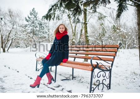 Blonde girl in red scarf and coat sitting at bench on winter day.