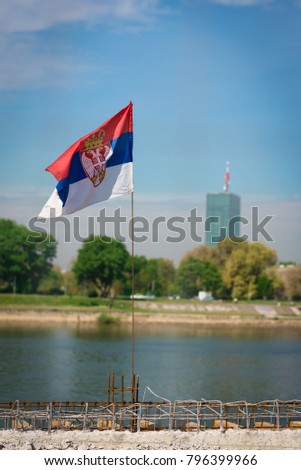 Serbian flag, on a rusty, steel wire rod, flying in the wind on a building site by the river Sava in Belgrade, with a building and a park in the background