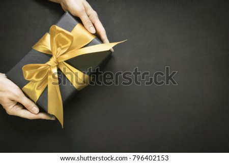 Gift box wrapped in black paper with gold ribbon in female hand on black surface. Copy space. Top view.