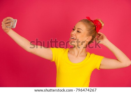 Beautiful young blond teenage woman in yellow t shirt  taking selfie over pink background.