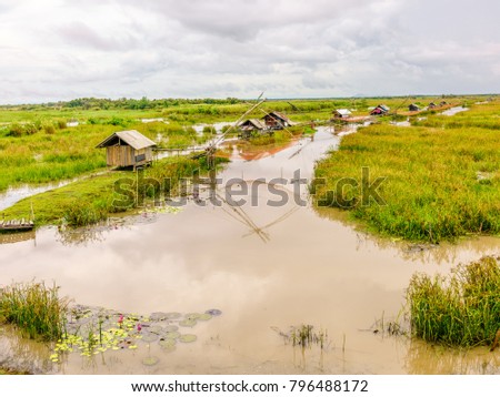 View from afar of fishermen huts along a small canal with their Lift Nets used to catch fishes in a blur background of flooded green fields and canal on horizon of Pathalung province, Thailand