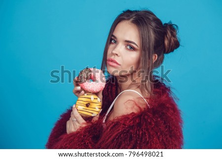 Attractive young woman in bright fur coat posing with donuts in hands.