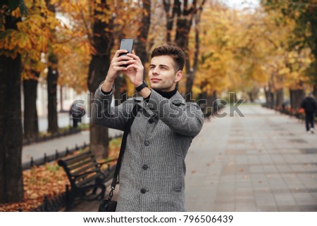 Handsome man in coat taking photo of beautiful autumn trees using his modern cellphone, while walking in empty park