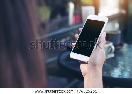 Mockup image of a woman's hand holding white mobile phone with blank black desktop screen and coffee cup in cafe