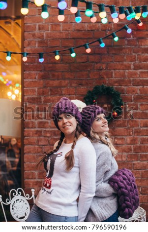 Two women in purple knitted hats, purple infinity scarf and blue jeans make posing with brick wall and Christmas garland on the background