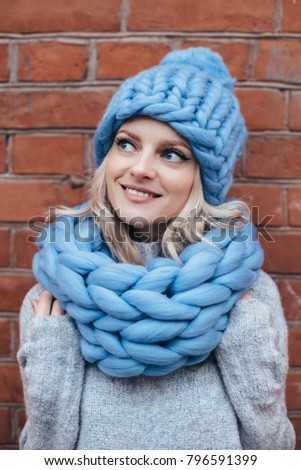 Young blonde woman in blue knitted hat, blue khitted infinity scarf and grey sweater make posing with brick wall on the background