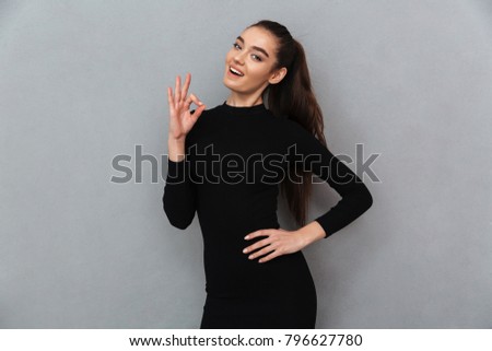 Picture of cheerful young beautiful woman standing over grey wall showing ok gesture. Looking camera.