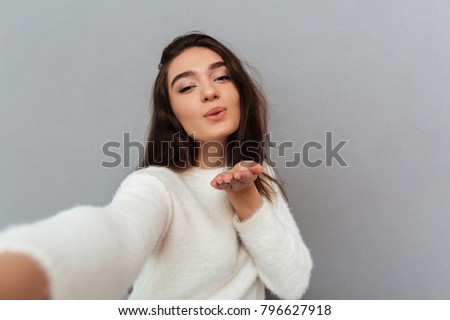 Close-up portrait of joyful brunette woman sending air kiss while taking selfie on mobile phone, isolated over gray background